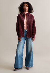 Corduroy overhemdjas in bordeauxrood met een borstzak, gedragen over een lichtroze coltrui, gecombineerd met broek met wijde pijpen in blauw en grijze schoenen.