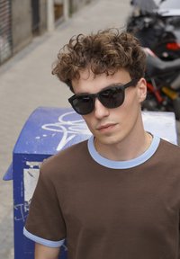 Pruun T-särk hele sinise krae ja varrukate detailidega; inimene mustade päikeseprillidega, seisab sinise postkasti juures tänaval.