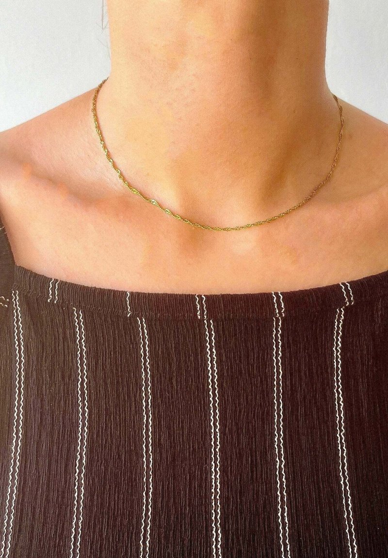 Gold-tone twisted necklace resting on bare skin, paired with a dark striped top featuring white accents. Simple and delicate design.
