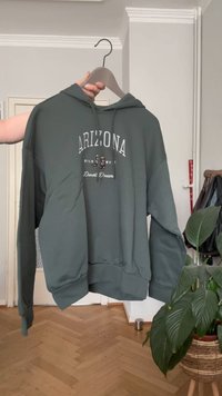 Zöld kapucnis felső egy fogason, rajta az "Arizona Wild West Desert Dreams" felirattal, egy kéz tartja egy szobában, amelynek fával burkolt a padlója és növények vannak benne.