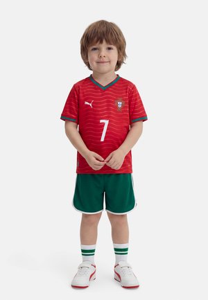 Niño pequeño de pie vistiendo una camiseta de fútbol roja de Portugal con el número 7, pantalones cortos verdes, calcetas blancas con rayas verdes y zapatillas blancas.