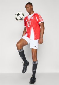 Rød og hvit Bayern München-drakt med logo, hvite shorts, svarte sokker med striper, svarte fotballsko, og en fotball i luften.