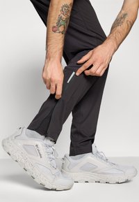 Lichtgrijze sportschoenen met een gevlekte zool, gedragen bij donkergrijze broek met een zijrits. Close-up van een hand die naar het ritsdetail wijst.