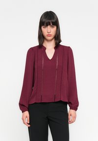Blouse bordeaux avec un décolleté en V, des manches bouffantes longues et un tissu texturé. Présente des coutures verticales et une coupe décontractée. Associée à un pantalon noir.
