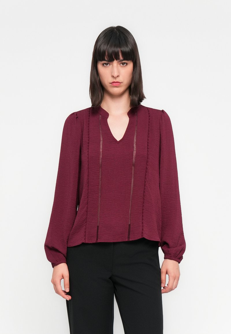 Blouse bordeaux avec un décolleté en V, des manches bouffantes longues et un tissu texturé. Présente des coutures verticales et une coupe décontractée. Associée à un pantalon noir.