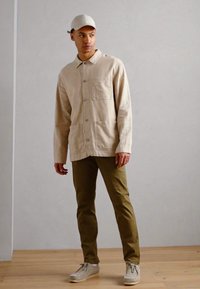 Cămașă din in bej cu nasturi și buzunar la piept, asortată cu pantaloni eleganți verzi măslinii și pantofi gri deschis. Prezintă de asemenea o șapcă gri deschis.