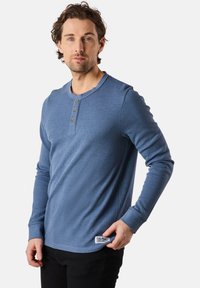 Blå långärmad henley-tröja med strukturerad tyg, funktioner en rundad halsringning, tre knappar och en etikett på den nedre vänstra sidan.
