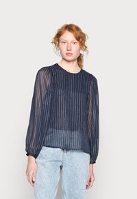Blusa azul-marinho, translúcida, com riscas verticais prateadas, mangas longas em balão e decote redondo. Usada com jeans azul claro.