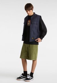 Marineblaues Stoffweste mit zwei vorderen Taschen, getragen über einem schwarzen Grafik-T-Shirt, kombiniert mit olivgrünen Cordshorts und schwarzen Turnschuhen.