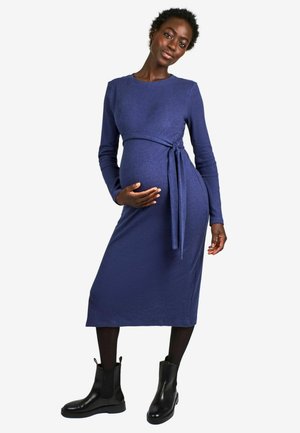 Schwangerschaftskleid aus dunkelblauem, geripptem Stoff, mit langen Ärmeln, taillierter Silhouette und einem Bindeband in der Taille. Kombiniert mit schwarzen Chelsea-Boots.