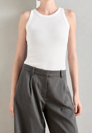 Person wearing a sleeveless white top tucked into high-waisted gray pinstripe trousers against a plain background.