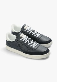 Schwarze Ledersneaker mit weißen Schnürsenkeln, ausgestattet mit einer strukturierten weißen Zunge und einem Totenkopf- und Knochen-Logo an der Seite. Gummisohle.