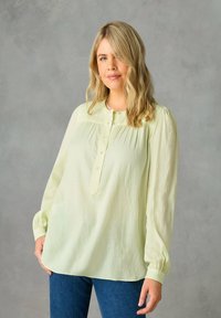 Blusa a maniche lunghe di colore giallo chiaro con chiusura a bottoni, dettaglio plissettato e polsini arricciati. Il tessuto appare morbido e leggero. Indossata con jeans blu.