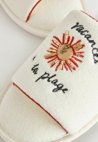 Zapatilla blanca de rizo con diseño bordado de sol decorada con cuentas doradas y texto en cursiva negra "vacances la plage."