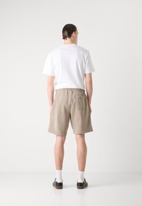 Pantaloni corti realizzati in tessuto beige con vita elasticizzata, dotati di una tasca posteriore. Abbinati a una maglietta bianca semplice e scarpe nere.