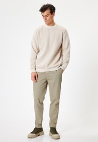 Beige sweatshirt med rund hals, parret med lyse khaki bukser og olivengrønne ankelstøvler. Har en subtil logo detalje på siden.