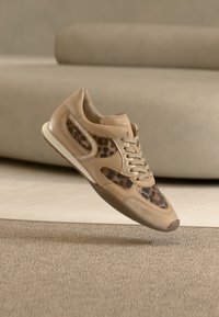 Μπεζ αθλητικό παπούτσι με πάνελ λεοπάρ, μπεζ κορδόνια και λευκή σόλα, αιωρούμενο πάνω από μια υφασμάτινη μπεζ μοκέτα σε ένα δωμάτιο με ουδέτερους τόνους.