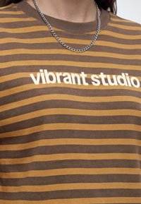Gros plan d'une personne portant une chemise rayée marron et moutarde avec un texte blanc "vibrant studio" et un collier chaîne argenté.