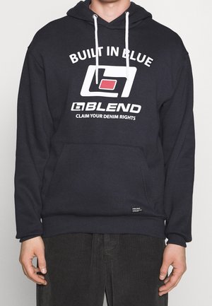 Personne portant un sweat à capuche noir avec des cordons blancs, arborant les textes et logos « Built in Blue », « Blend » et « Claim Your Denim Rights » sur le devant.