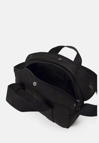 Czarna torba na ramię z tkaniny z regulowanym paskiem, otwartą zapinaną na zamek główną komorą, przednią kieszenią na zatrzask i górnym uchwytem na białym tle.