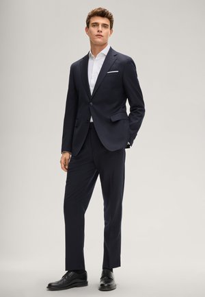 Mężczyzna stojący, ubrany w granatowy garnitur, białą koszulę, czarne eleganckie buty i białą poszetkę na jednolitym tle.