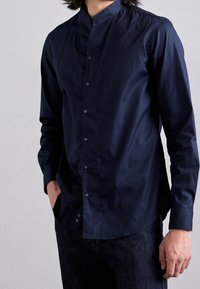 Marineblaue Langarmbluse mit Stehkragen, vorderem Knopfverschluss und glatter Textur. Kombiniert mit dunklen Jeans.