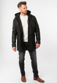 Zwarte leren jas met capuchon, geribbelde kraag, rits aan de voorkant en zijzakken, gedragen over een wit shirt en grijze spijkerbroek. Bruine laarzen.