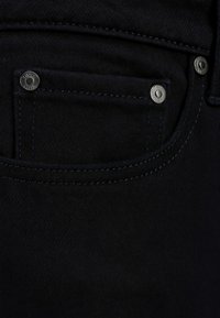 Jeans in denim nero con una piccola tasca frontale dotata di un bottone in metallo argentato, contrapposti da cuciture blu scuro lungo le cuciture.