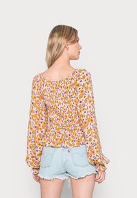 Blusa floral com padrões em amarelo, laranja e rosa; mangas franzidas; corte solto; tecido leve; combinada com calções de ganga rasgados.