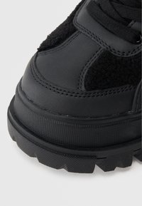 Fekete sportcipő, amely sima bőr és texturált szövet kombinációjából áll. Masszív gumi talp mintázott tapadással. Lekerekített orr kialakítás.