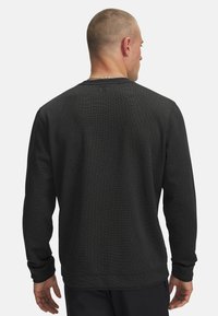 Mann mit kurzen Haaren, der ein schwarzes, strukturiertes Langarmshirt trägt, von hinten gegen einen schlichten Hintergrund betrachtet.