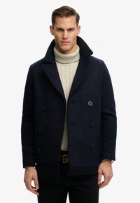 Man met een marineblauw dubbelgeknoopt pea-coat over een beige kabelgebreide coltrui, staand tegen een effen witte achtergrond.