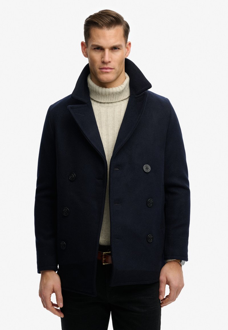 Man met een marineblauw dubbelgeknoopt pea-coat over een beige kabelgebreide coltrui, staand tegen een effen witte achtergrond.