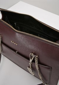 Bordová kožená kabelka s hladkou texturou, zlatým kováním, zapínáním na zip a vnitřními kapsami. Má odnímatelný řetízkový popruh a detail s logem.