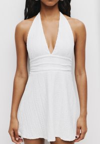 Weißes Halterkleid mit tiefem V-Ausschnitt, strukturiertem Stoff und gerafftem Taillenbereich. Kurzes, schwingendes Design mit dezenten Mustern.