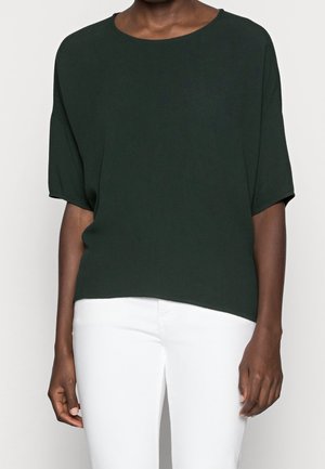 Personne portant un haut ample à manches courtes vert foncé associé à un pantalon blanc ajusté, visible du cou aux hanches sur un fond neutre.