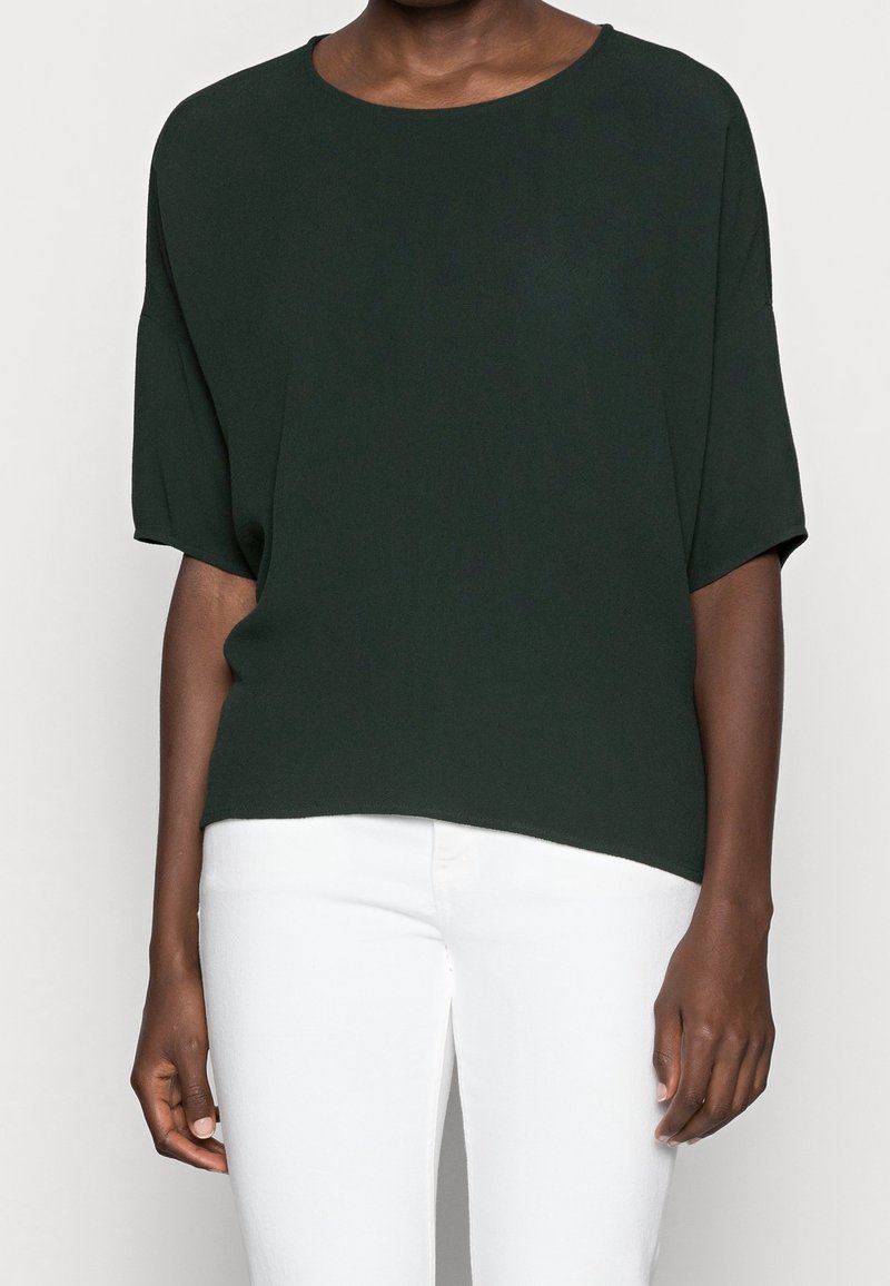 Personne portant un haut ample à manches courtes vert foncé associé à un pantalon blanc ajusté, visible du cou aux hanches sur un fond neutre.