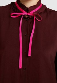 Blusa bordeaux con colletto plissettato, dotata di nastri rosa acceso e bottoni sul davanti. Tessuto dalla texture liscia.