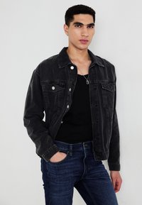 Chaqueta de mezclilla negra con botones plateados, dos bolsillos en el pecho y un corte holgado, combinada con una camiseta negra sin mangas y jeans azules.