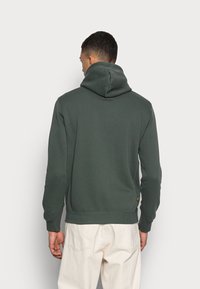 Haut à capuche vert avec cordon, poignets et ourlet côtelés, texture lisse et petite étiquette logo à la taille.