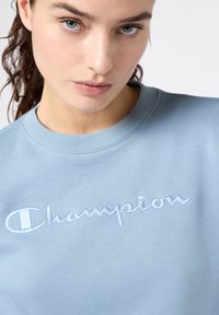 Felpa azzurra chiara realizzata in tessuto morbido, con logo "Champion" ricamato di un blu leggermente più scuro. Collo rotondo classico e polsini a coste.