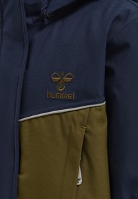 Marineblau und olivgrüne Jacke aus strukturiertem Material, mit einem Logo-Patch, Reißverschluss und kontrastierendem weißem Saum an der Schulter.