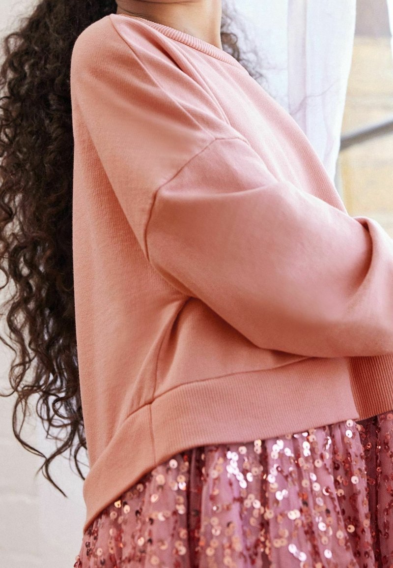 Sweat-shirt rose clair avec un col et des poignets côtelés, associé à une jupe rose à sequins avec un design en couches et une texture scintillante.