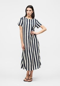 Γυναίκα φοράει μια μαύρη και λευκή κάθετα ριγωτή μίντι φόρεμα με κοντά μανίκια, στέκεται με το ένα χέρι στη μέση, ξυπόλητη με σανδάλια κοντά.