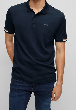 Mann trägt ein marineblaues Kurzarm-Poloshirt mit "BOSS" Logo auf der Brust und gestreiften Abschlüssen an den Ärmeln, kombiniert mit dunkelblauen Jeans.