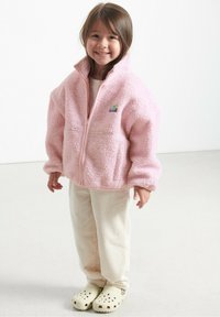 Chaqueta de forro polar rosa claro con cuello alto, cierre de cremallera y parche de logo. Combinada con pantalones de pana color crema y zuecos amarillo claro.