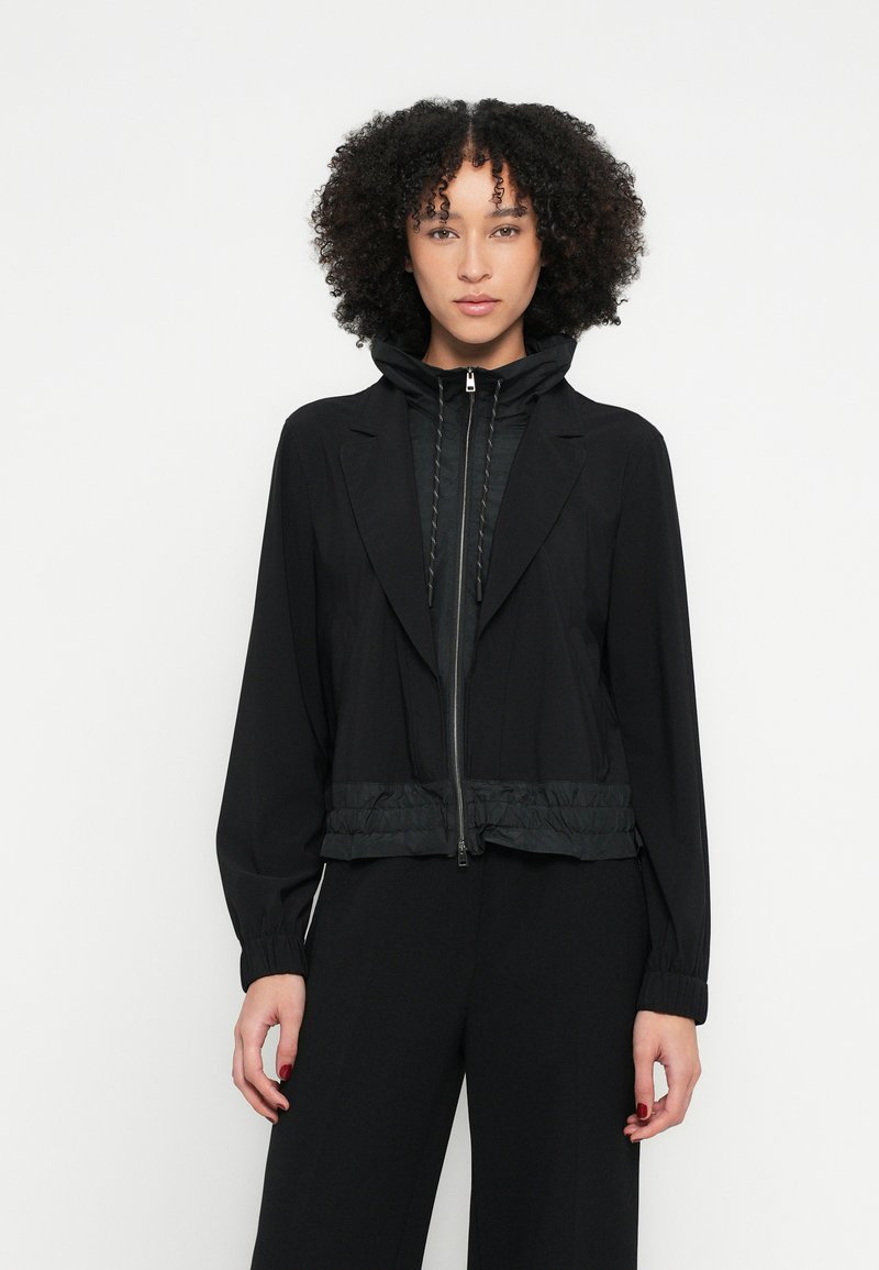 Jachetă neagră cu fermoar frontal, guler structurat și bandă elastică în talie. Are o glugă și manșete ribbed, realizată dintr-un material neted.