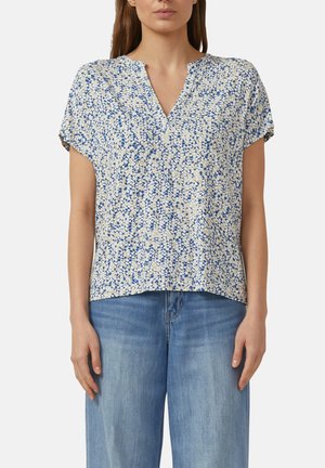 Kobieta ubrana w bluzkę z krótkim rękawem, w niebiesko-beżowy kwiatowy wzór z dekoltem w serek, zestawioną z jasnoniebieskimi dżinsami.