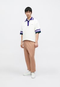 Biała koszulka polo z fioletowym kołnierzykiem i pasiastymi rękawami, beżowe spodnie plisowane i białe tenisówki, prezentowane na męskim modelu.