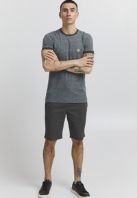 T-shirt gris à manches courtes avec des bords noirs, associé à un short sombre. Le modèle a également des tatouages visibles et porte des baskets noires.
